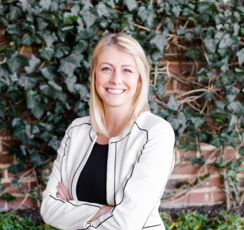 Noelle McHugh headshot, woman smiling in front of vine grass wall